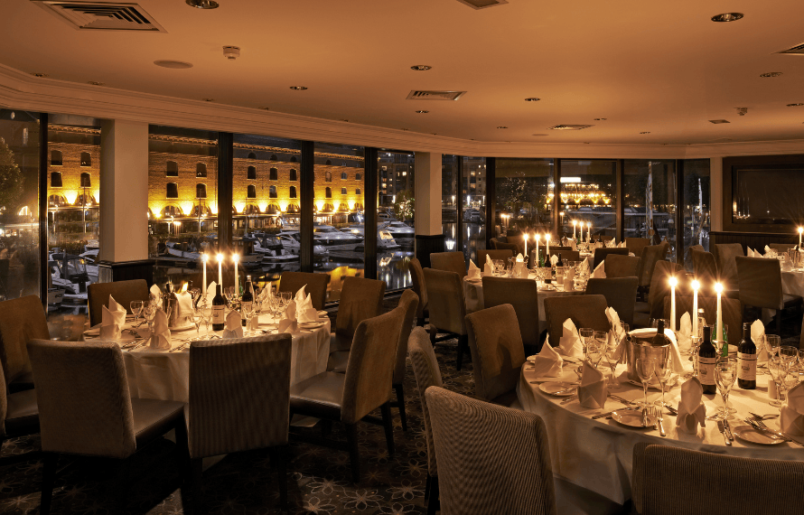 Elegant dining room with candlelit tables set for an evening event, overlooking lit buildings and water through large windows